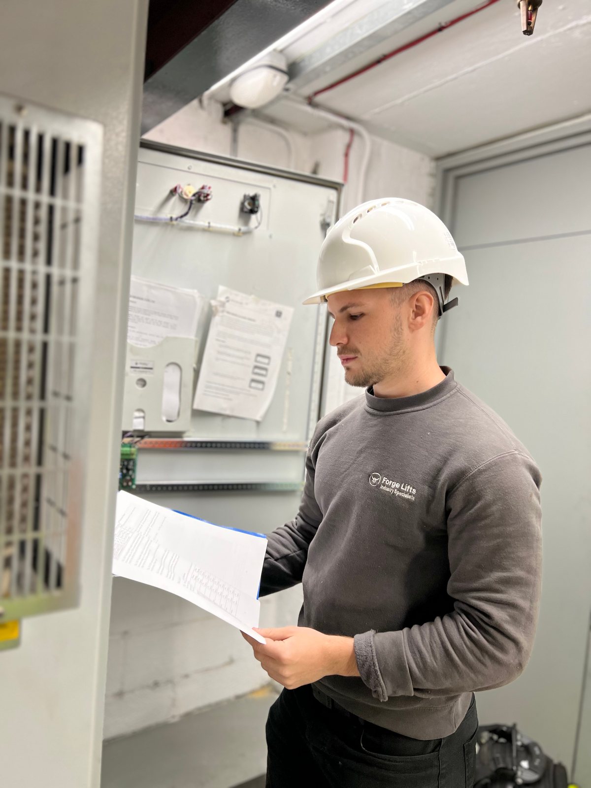 Forge Lifts engineer in a hard hat reviewing documentation beside a lift control cabinet